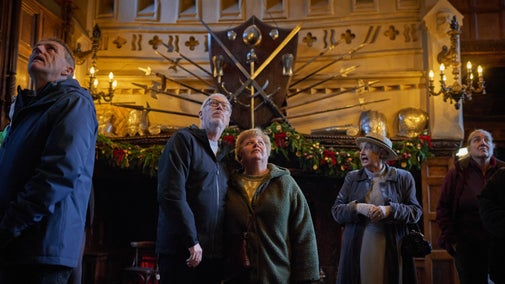 A couple stood in the centre of a Great Hall looking up in front of a mounted display of swords.